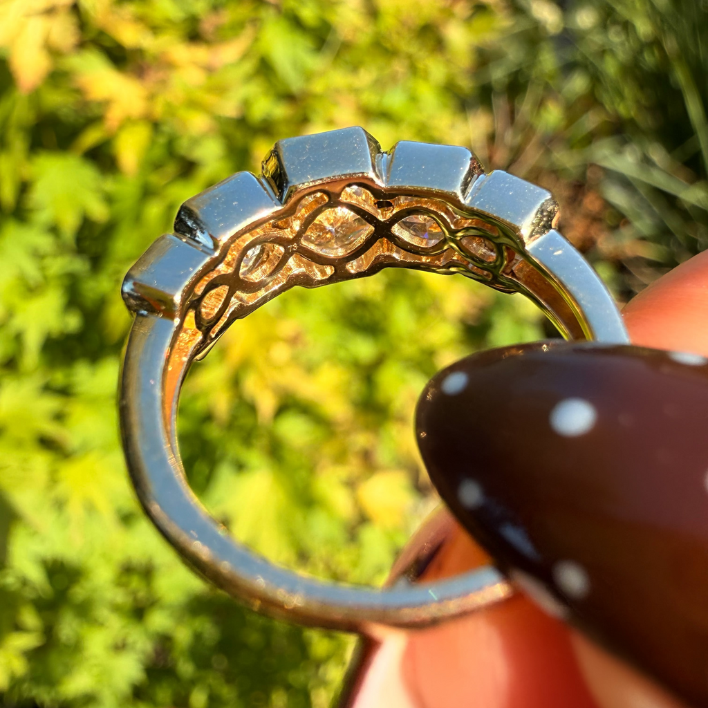 A close-up of a silver ring held by a hand, showing a decorative openwork band with a central oval cutout, set against a blurred green outdoor background.