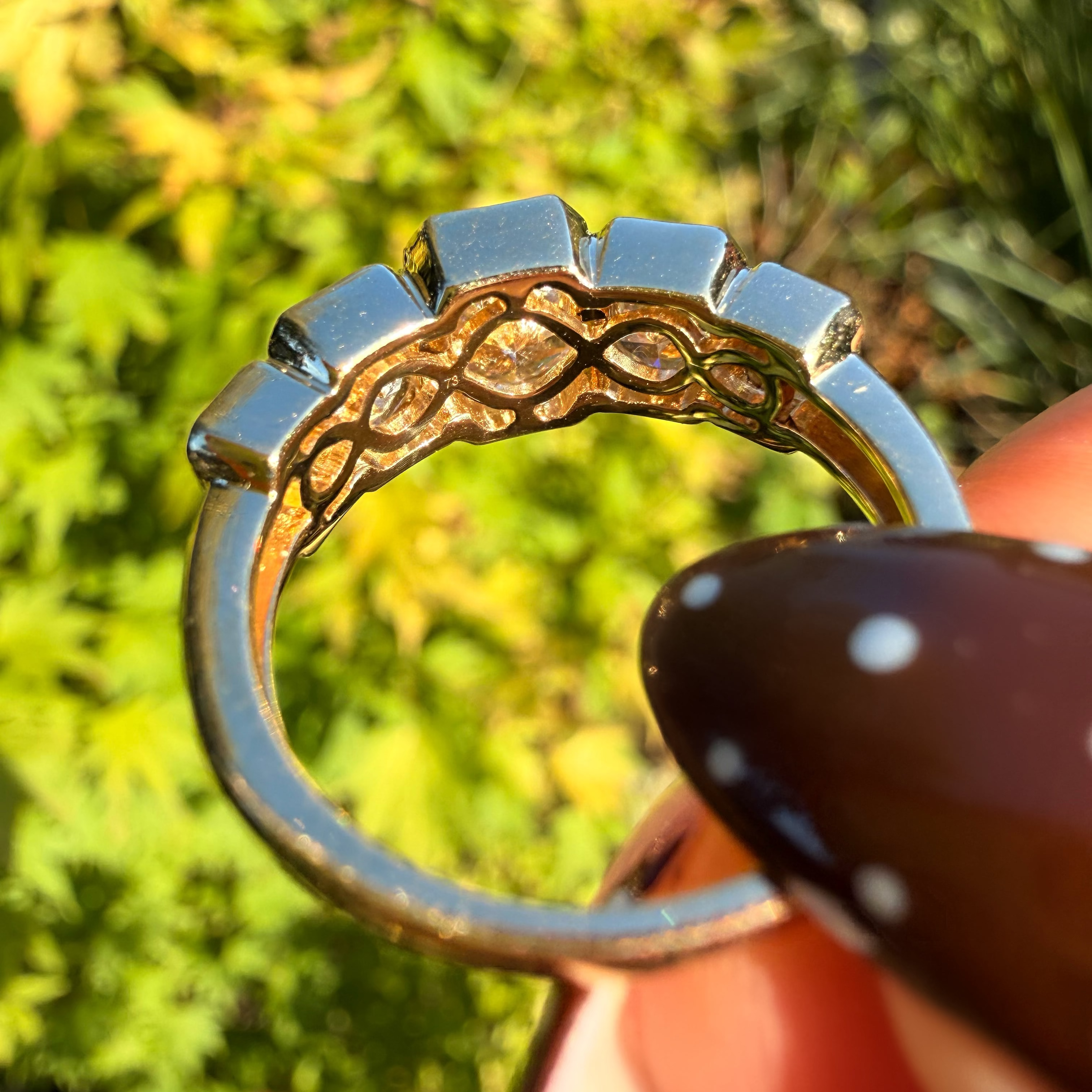 A close-up of a silver ring held by a hand, showing a decorative openwork band with a central oval cutout, set against a blurred green outdoor background.