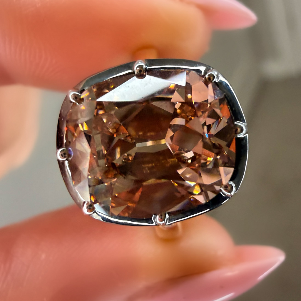 Brown gemstone ring held by a hand with pink nail polish against a neutral background