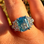 A close-up of a ring worn on a hand outdoors. The ring features a large cushion-cut blue gemstone with smaller clear stones on either side, set in a silver-toned metal.