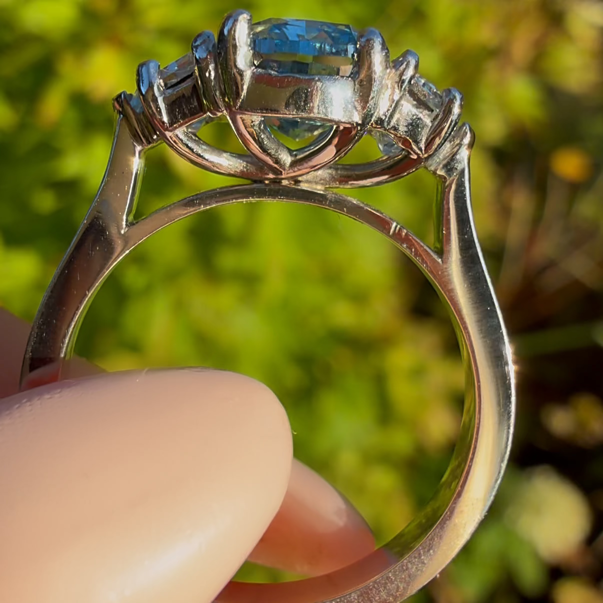 A close-up of a silver ring with a rectangular blue gemstone in a prong setting, held between fingers against a blurred green outdoor background.