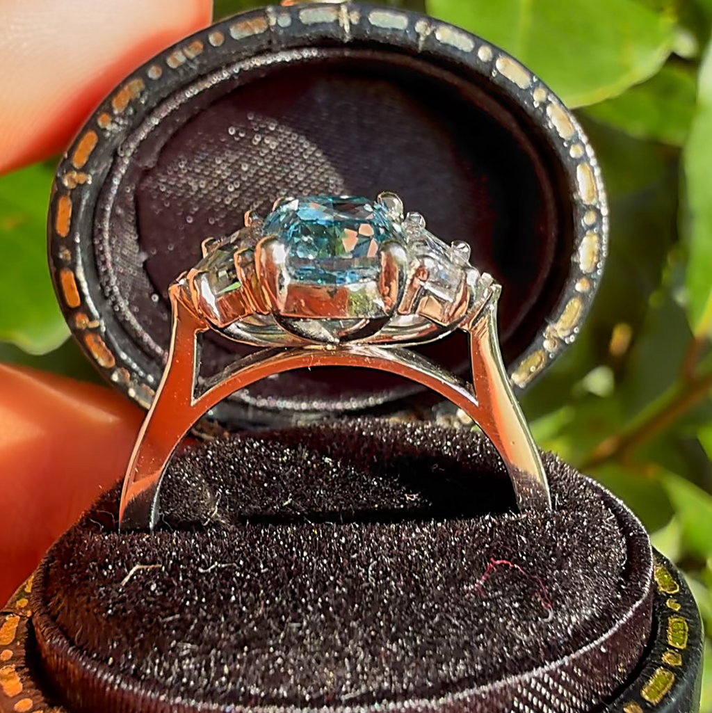 A silver-toned ring with a blue-green marquise-cut gemstone and smaller side stones, displayed in an open black ring box held by a hand against a background of green leaves.