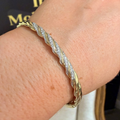 A close-up of a gold-toned twisted rope bangle bracelet with pavé-set clear stones, worn on a person's wrist. A matching gold ring with a clear gemstone is also visible on the hand.