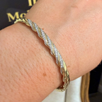A close-up of a gold-toned twisted rope bangle bracelet with pavé-set clear stones, worn on a person's wrist. A matching gold ring with a clear gemstone is also visible on the hand.