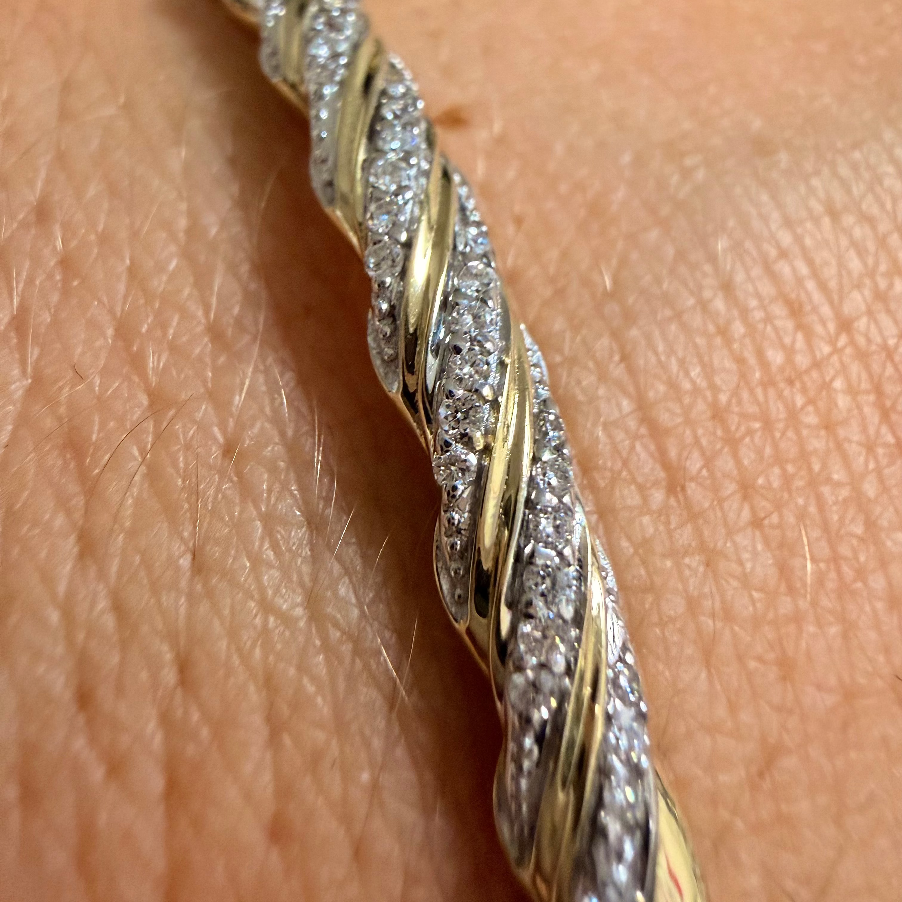 A close-up of a twisted rope-style bracelet worn on a person's wrist. The bracelet features alternating polished yellow gold and pavé-set clear stones, with the skin and a blurred dark jewelry box visible in the background.