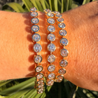 Close-up of a wrist wearing two gold bracelets with clear stones against a green leafy background