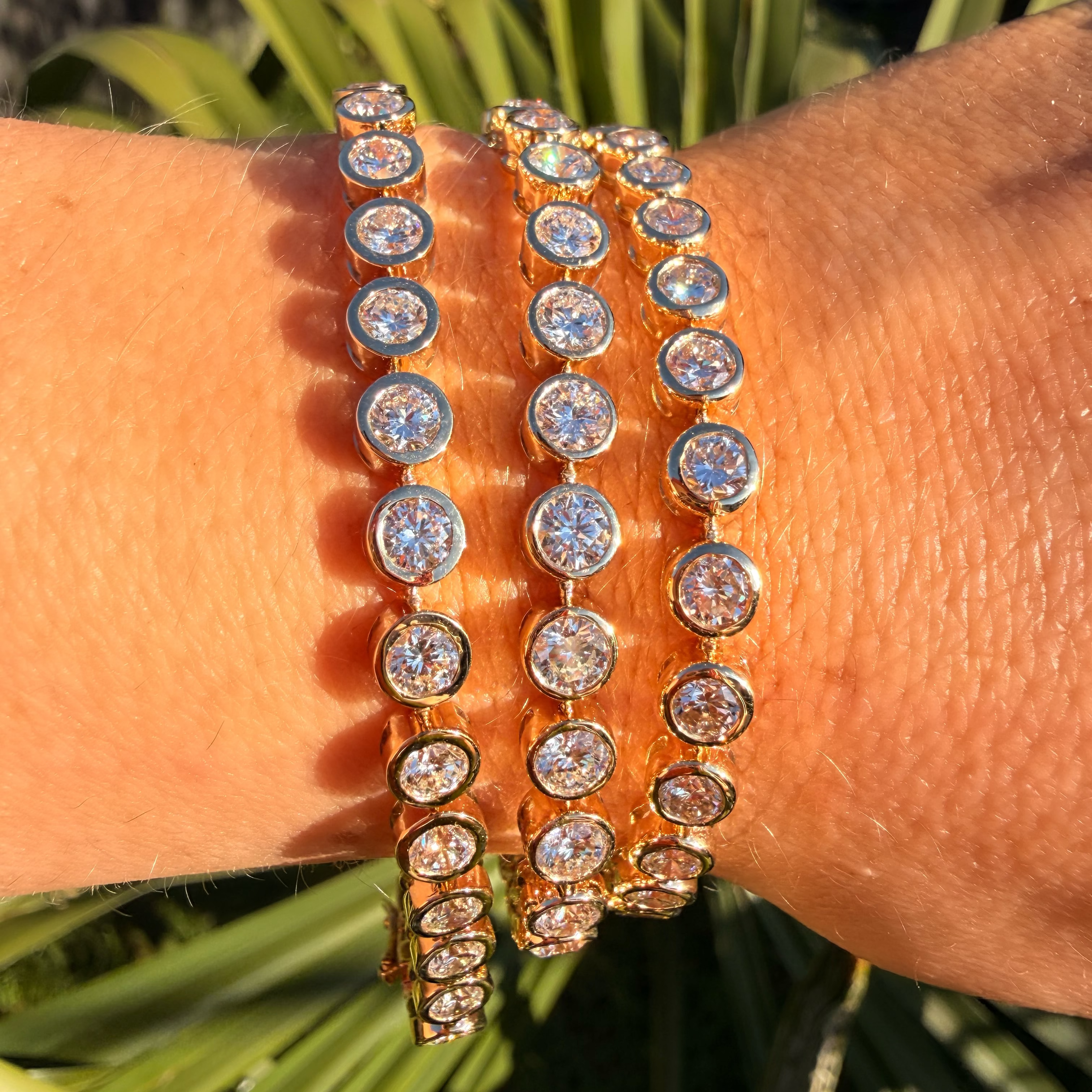 Close-up of a wrist wearing two gold bracelets with clear stones against a green leafy background