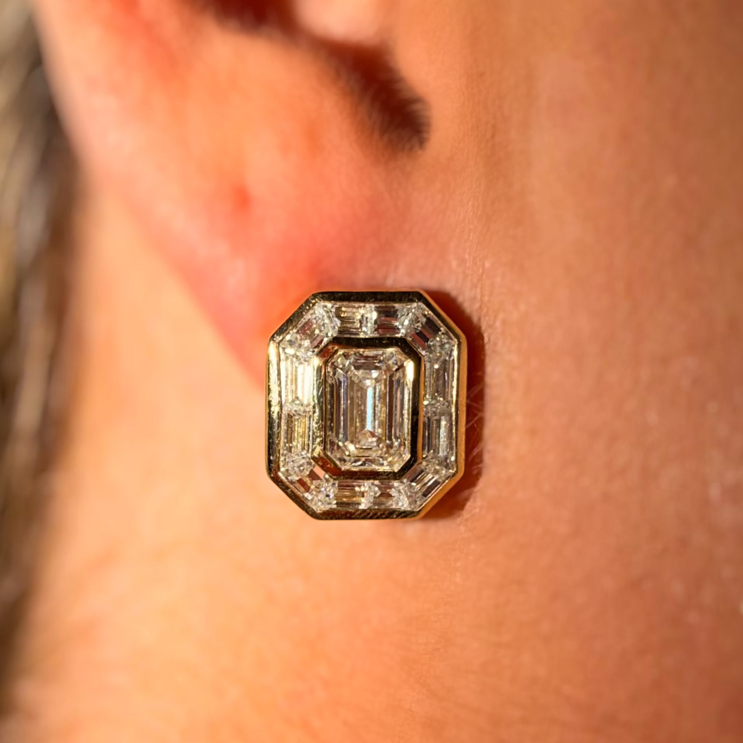 A close-up of a person's ear wearing a gold-toned geometric stud earring with a central emerald-cut clear gemstone and smaller surrounding stones.