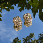 A pair of gold-toned statement earrings with large emerald-cut clear gemstones, shown floating against a reflective blue sky with green tree leaves reflected in the surface below.