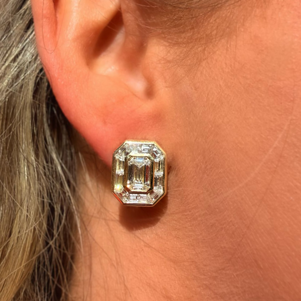 A close-up of a person's ear wearing a gold stud earring with an emerald-cut clear gemstone.