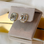 A pair of gold-toned stud earrings with emerald-cut clear stones, displayed on a beige velvet jewelry box.