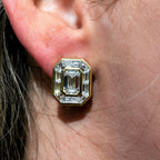 A close-up of a person's ear wearing a gold-toned stud earring with a large emerald-cut clear gemstone.