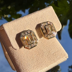 A pair of gold-toned stud earrings with large emerald-cut clear gemstones, displayed on a beige velvet jewelry box with an outdoor sky and tree reflection in the background.