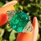 A close-up of a ring held between fingers, featuring a large emerald-cut green center stone with clear side stones in a silver-toned setting, against a blurred green outdoor background.