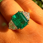 A close-up of a ring worn on a finger outdoors, featuring a large rectangular green emerald-cut center stone with two clear triangular side stones and a gold-toned setting.