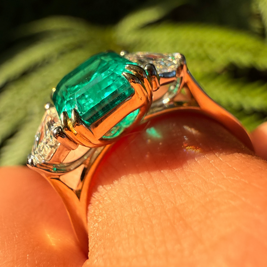 A close-up shot of a ring worn on a finger, featuring a large emerald-cut green center stone with smaller clear stones on either side, set in a warm-toned metal against a blurred green background.