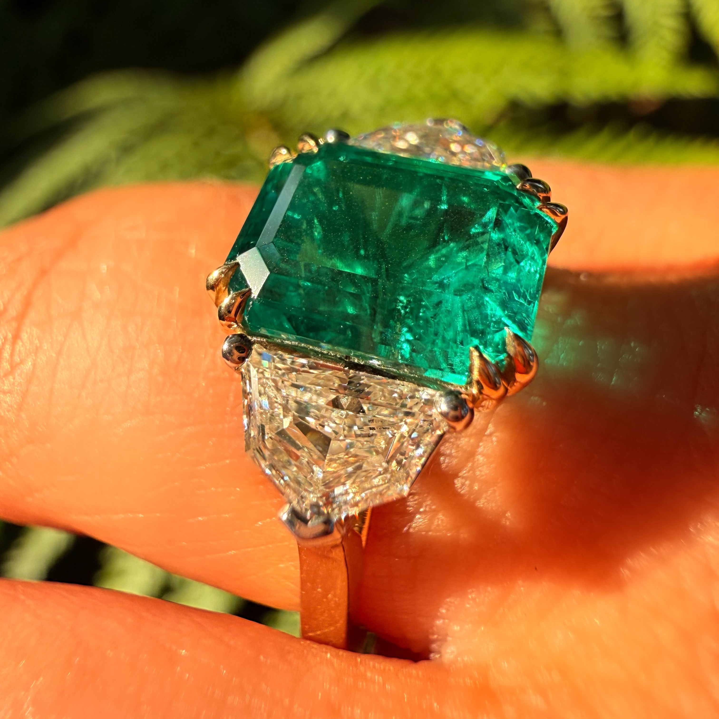 A close-up of a ring worn on a finger, featuring a large emerald-cut green gemstone in a gold setting with diamond accents below, against a blurred green background.