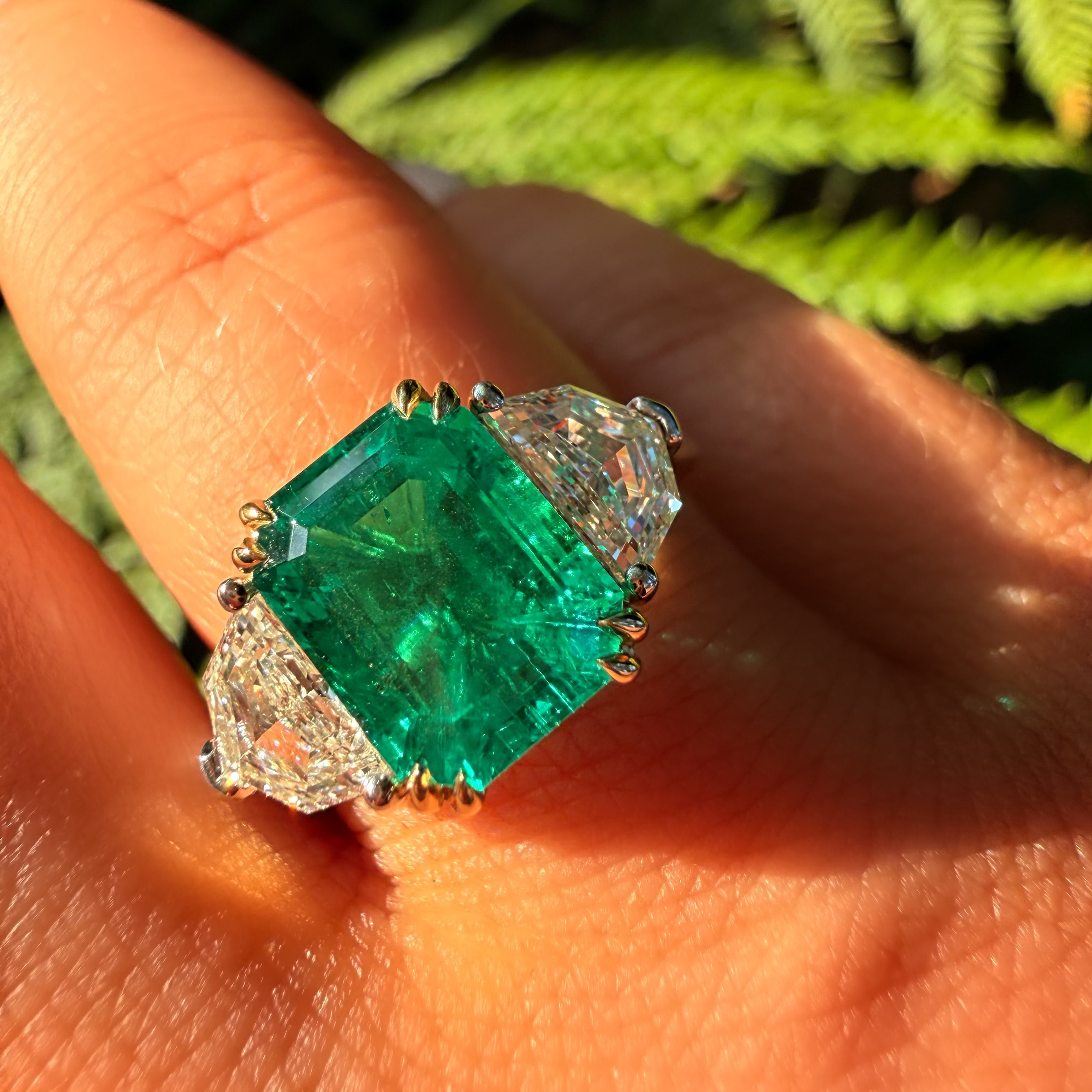 A close-up of a hand wearing a ring with a large emerald-cut green center stone and clear side stones in a gold-toned setting, with blurred green foliage in the background.