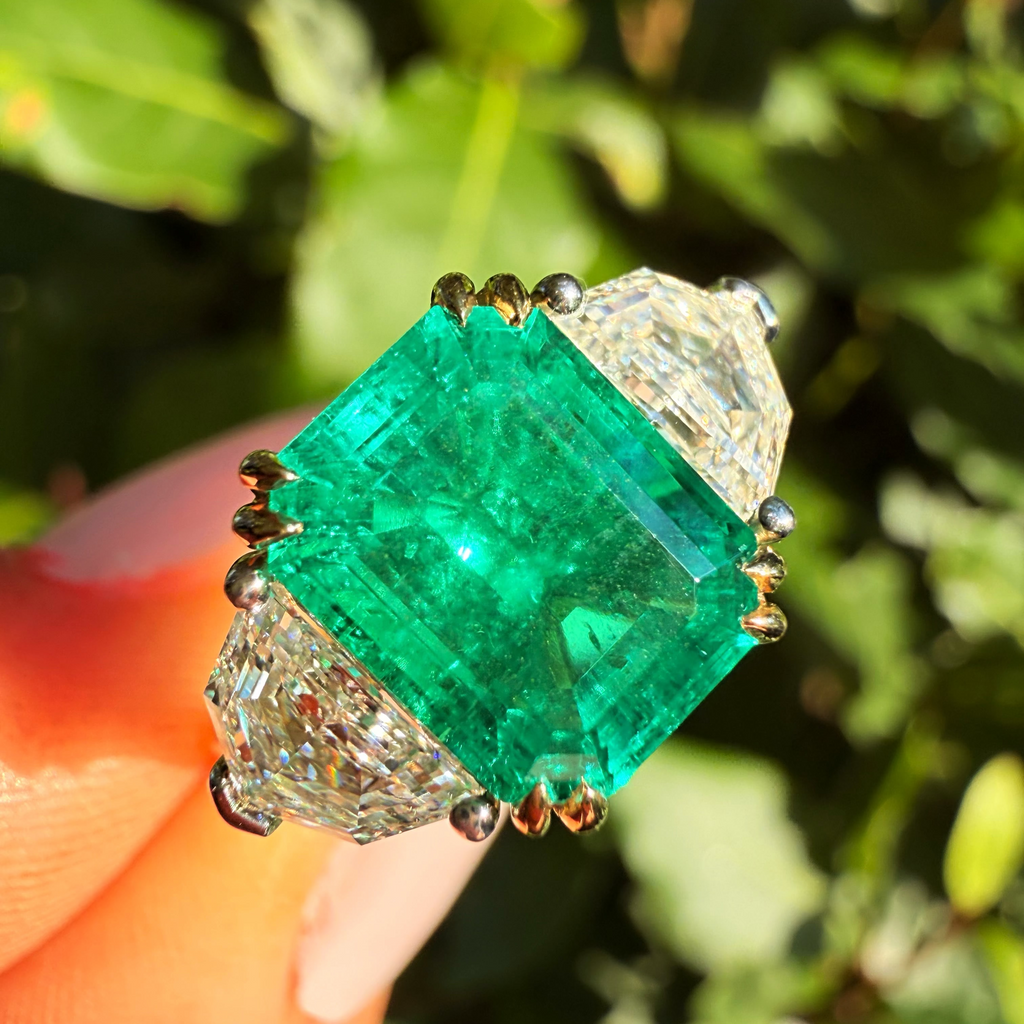 A close-up of a ring held on a finger, featuring a large emerald-cut green center stone with clear side stones in a gold-toned setting, against a blurred green outdoor background.