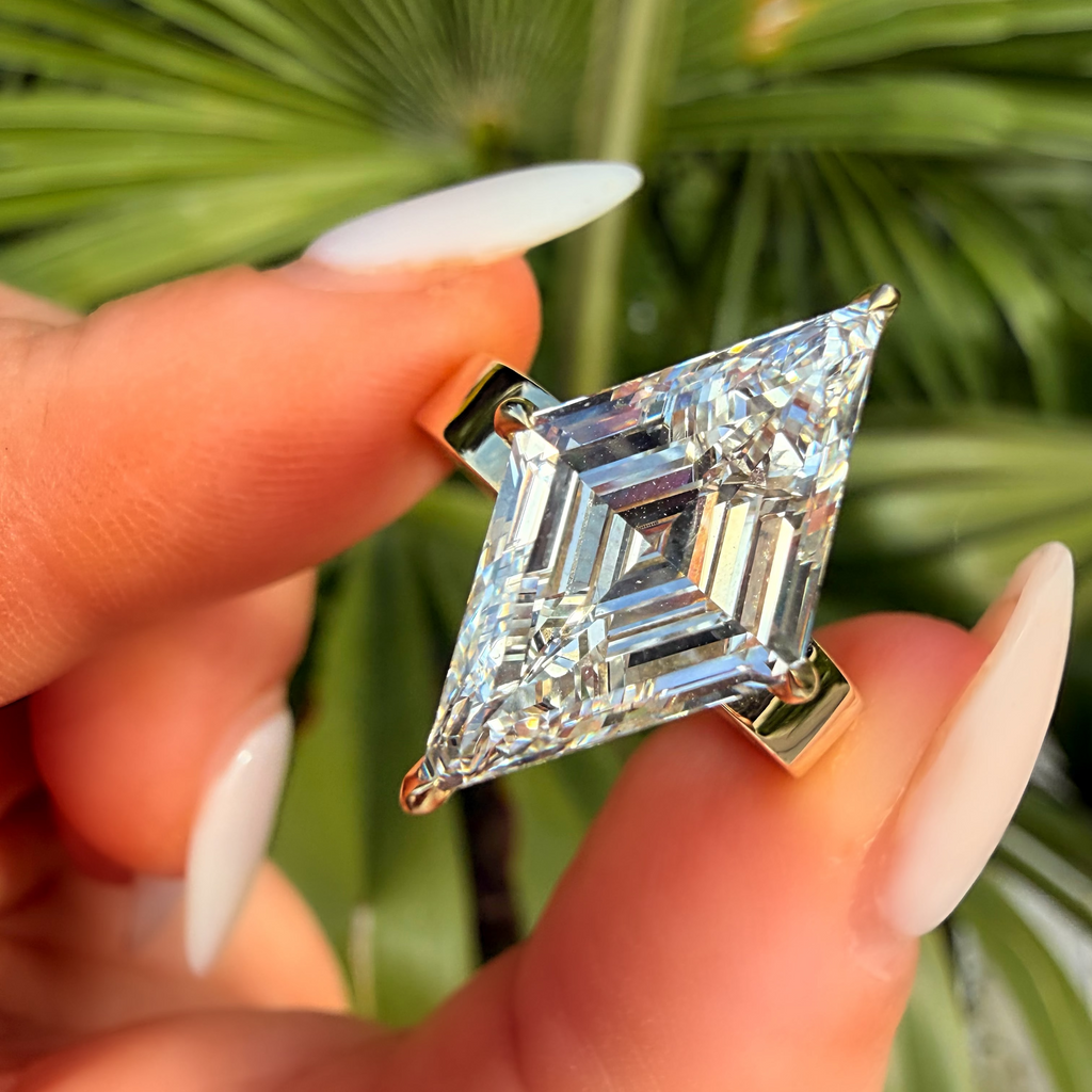A close-up of a hand with long white manicured nails holding a gold ring set with a large brilliant-cut diamond. The background is a soft-focus green palm leaf.