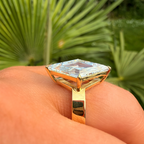 A gold ring with a large cushion-cut clear gemstone worn on a hand, with a blurred background of green palm leaves.