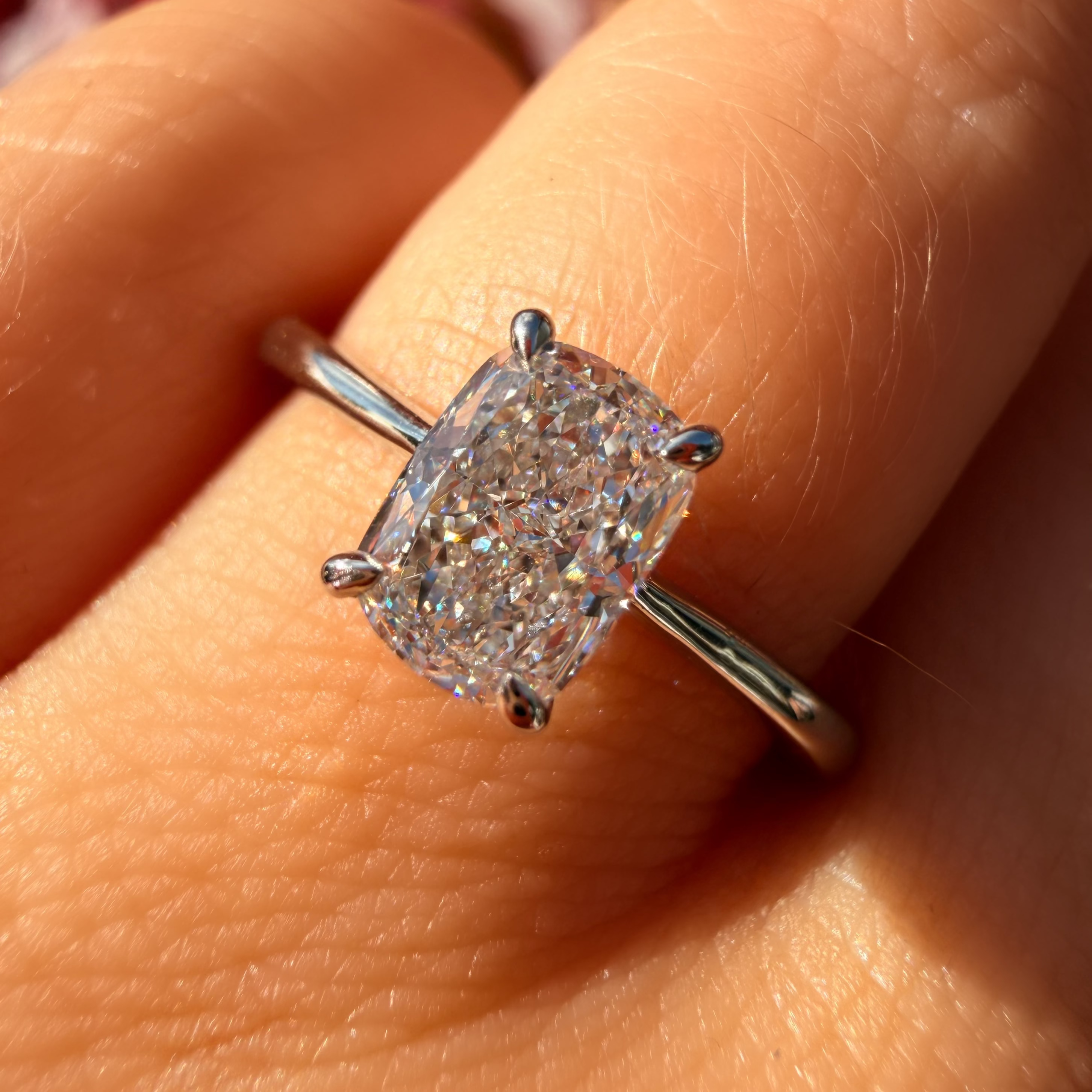 A close-up of a silver ring with a cushion-cut clear gemstone worn on a finger. The ring is shown in bright natural light with a blurred background.