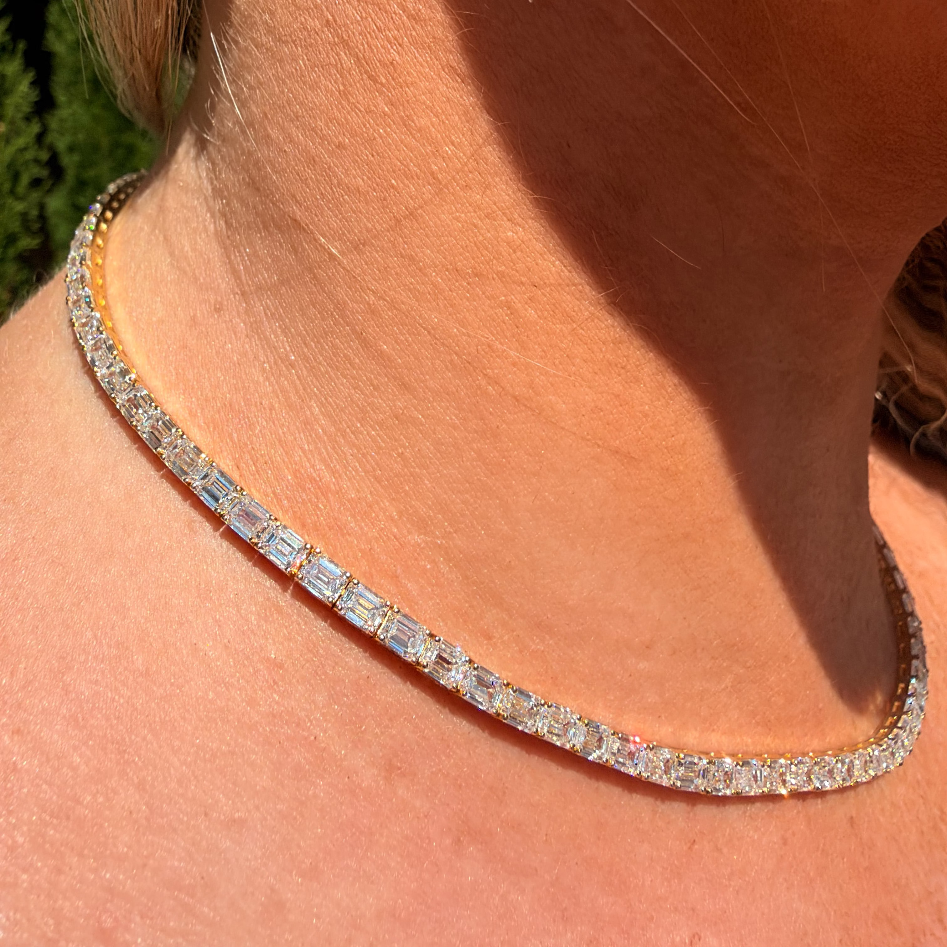 A close-up of a woman's neck and collarbone wearing a diamond tennis necklace with a gold setting, alongside a matching drop earring.