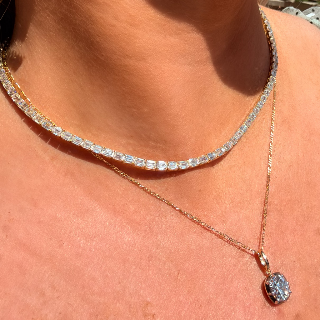 A close-up of a woman's neck and chest wearing a layered necklace set. The top layer is a tennis-style choker with clear rectangular gemstones in a silver-toned setting, and the bottom layer is a delicate rose gold chain with a small oval clear gemstone pendant.