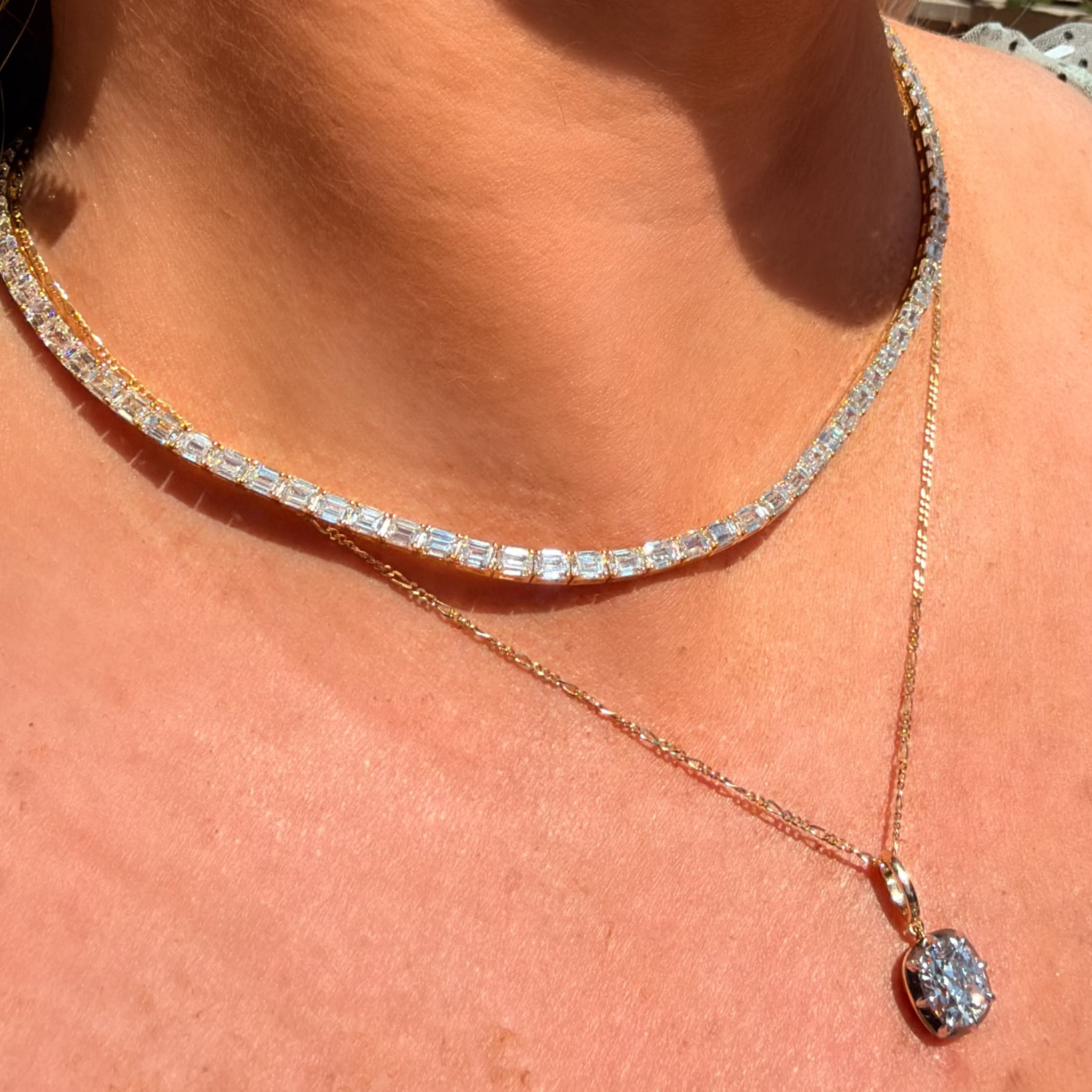 A close-up of a woman's neck and chest wearing a layered necklace set. The top layer is a tennis-style choker with clear rectangular gemstones in a silver-toned setting, and the bottom layer is a delicate rose gold chain with a small oval clear gemstone pendant.