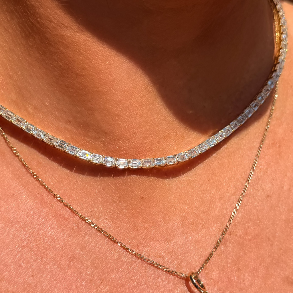 A close-up of a person's neck and chest wearing two layered necklaces: a silver tennis-style chain necklace and a gold chain necklace with a round clear gemstone pendant.