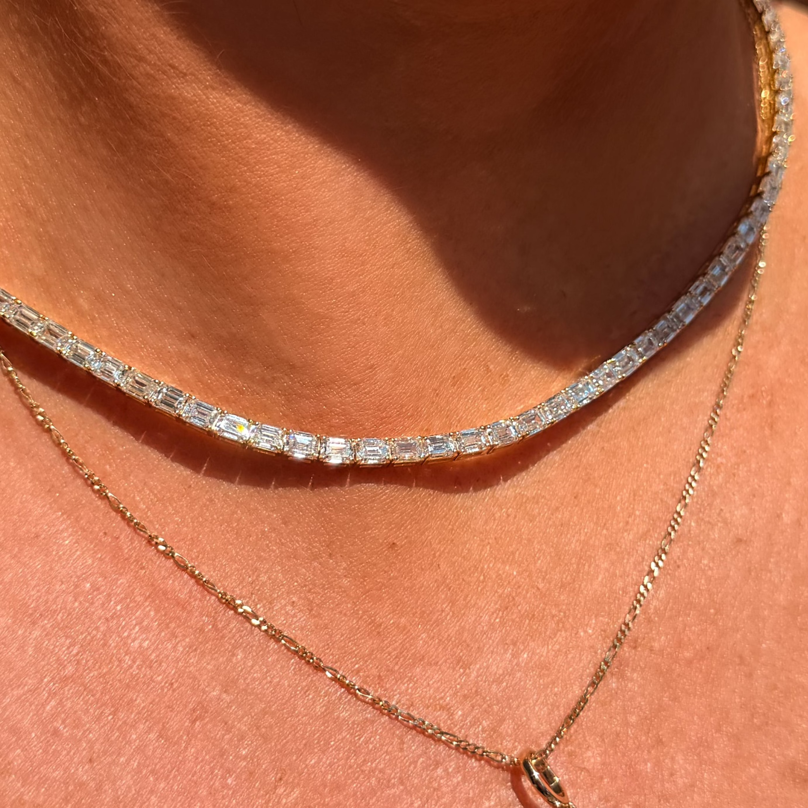 A close-up of a person's neck and chest wearing two layered necklaces: a silver tennis-style chain necklace and a gold chain necklace with a round clear gemstone pendant.