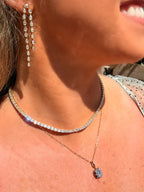 A close-up lifestyle shot of a woman wearing a layered jewelry set. The set includes a gold tennis necklace with clear stones, a delicate gold chain with a round clear stone pendant, and a matching gold drop earring.
