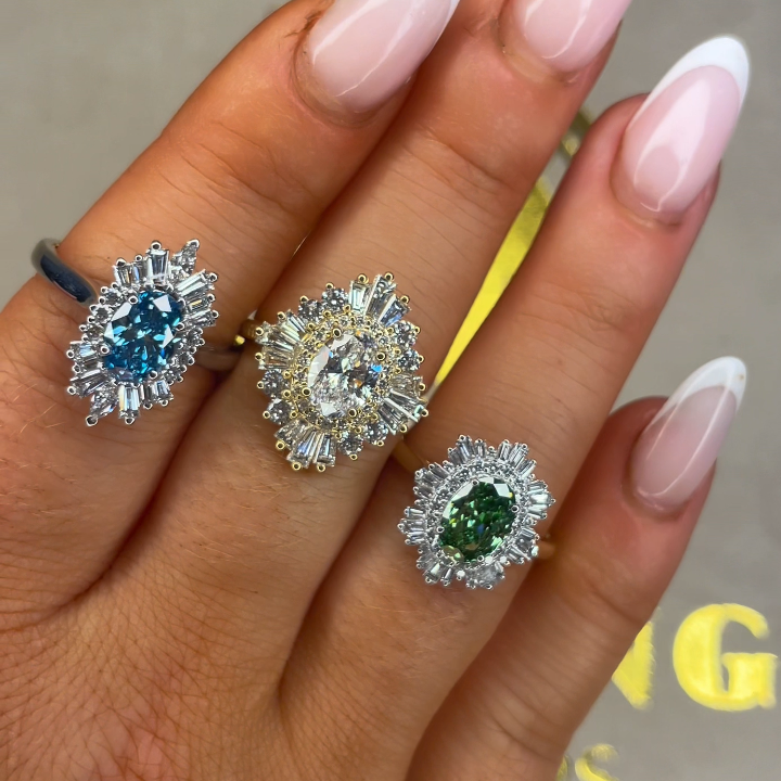 Hand wearing three diamond rings with different colored gemstones on a neutral background