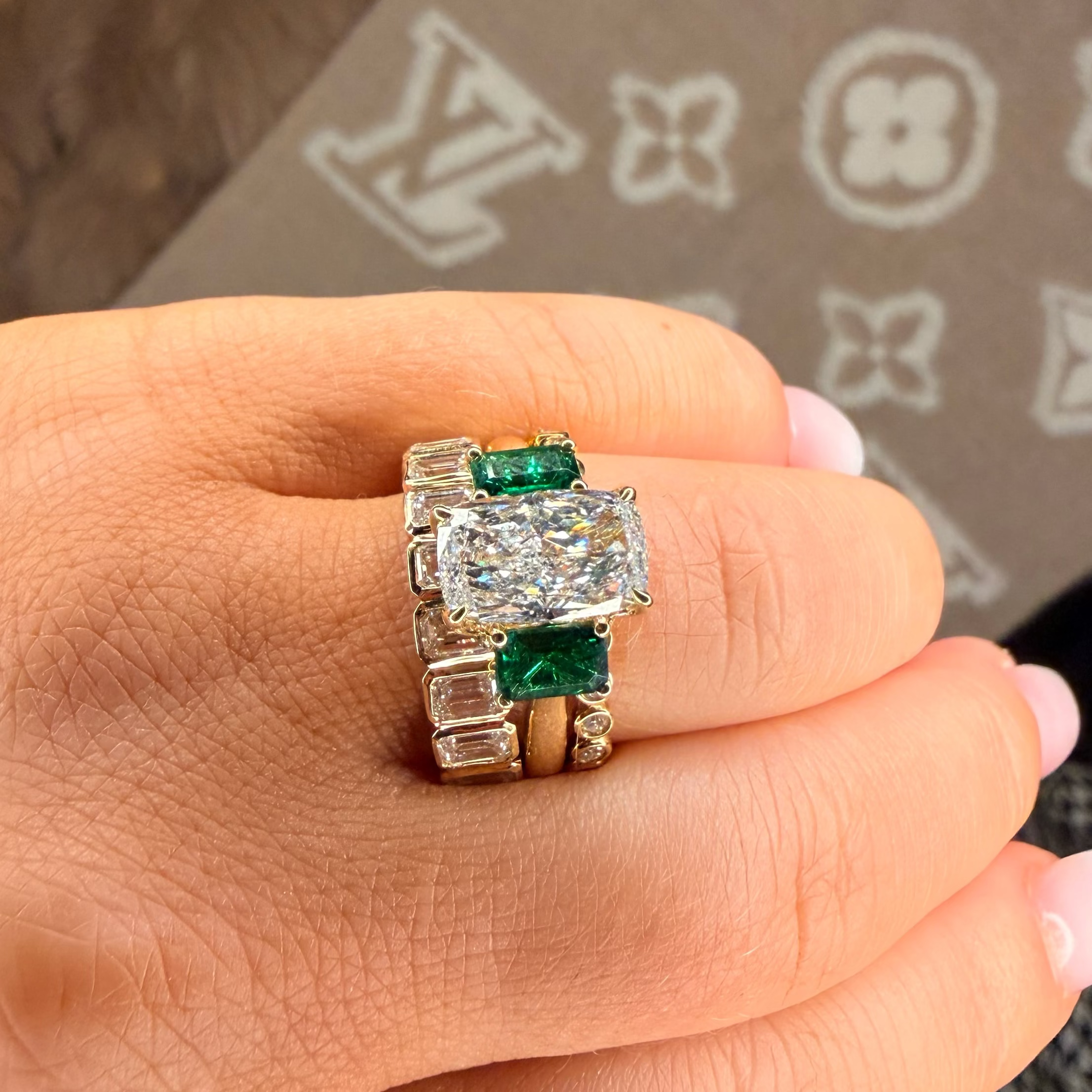 Gold ring with emeralds and diamonds on a hand, with a Louis Vuitton background.