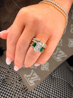 Close-up of a hand wearing a ring with emeralds and diamonds on a patterned surface.