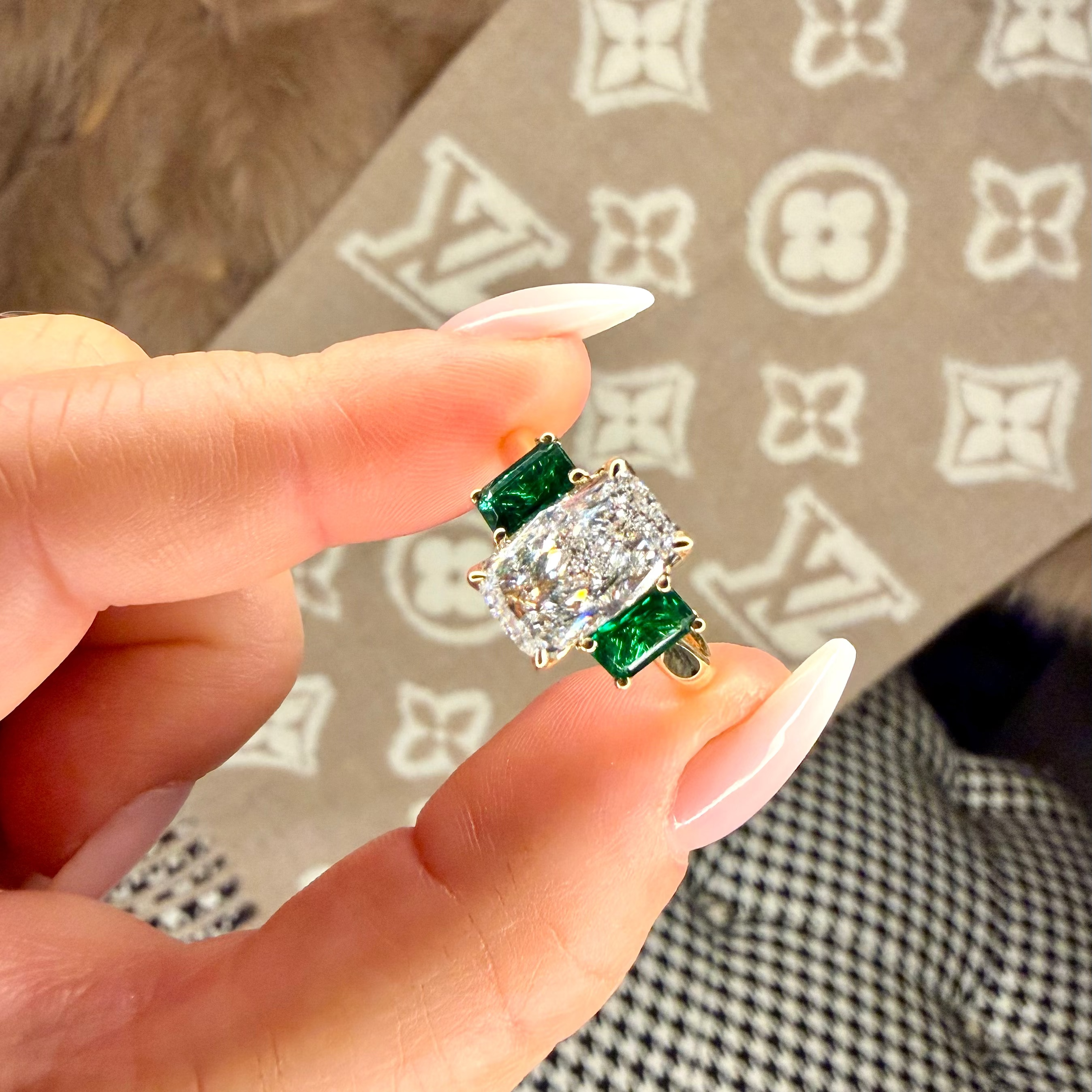 Hand holding a diamond ring with green gemstones against a patterned background