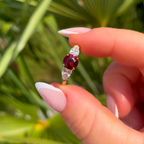Hand holding a ring with a red gemstone against a green leafy background