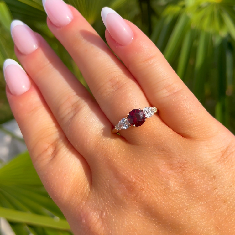 Hand wearing a ring with a red gemstone and diamond accents against a green leafy background