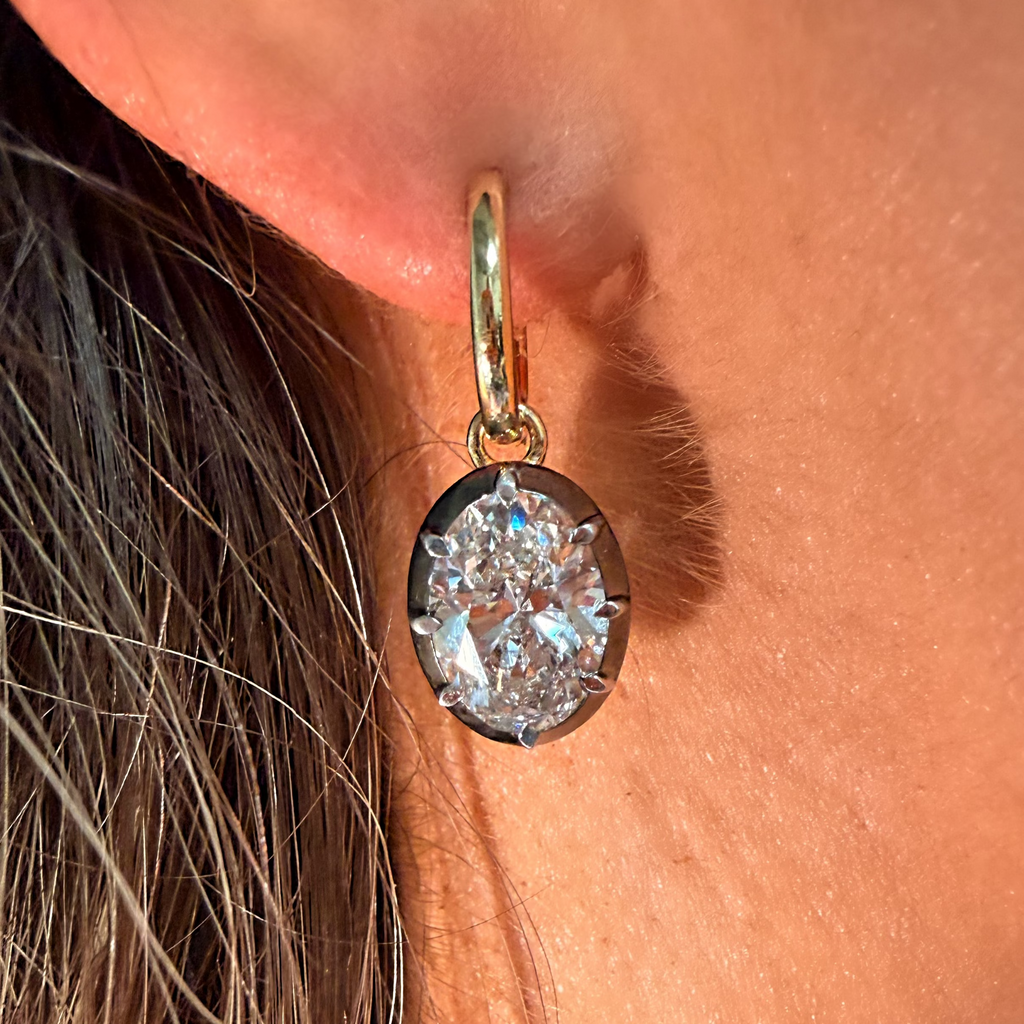 A close-up of a person's ear wearing a gold hoop earring with a round brilliant-cut clear gemstone pendant.