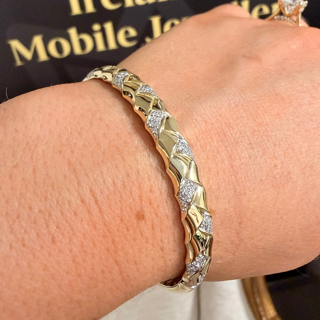 A close-up of a gold and silver pavé bangle bracelet worn on a person's wrist, with a ring visible on another hand and a dark jewelry box in the background.