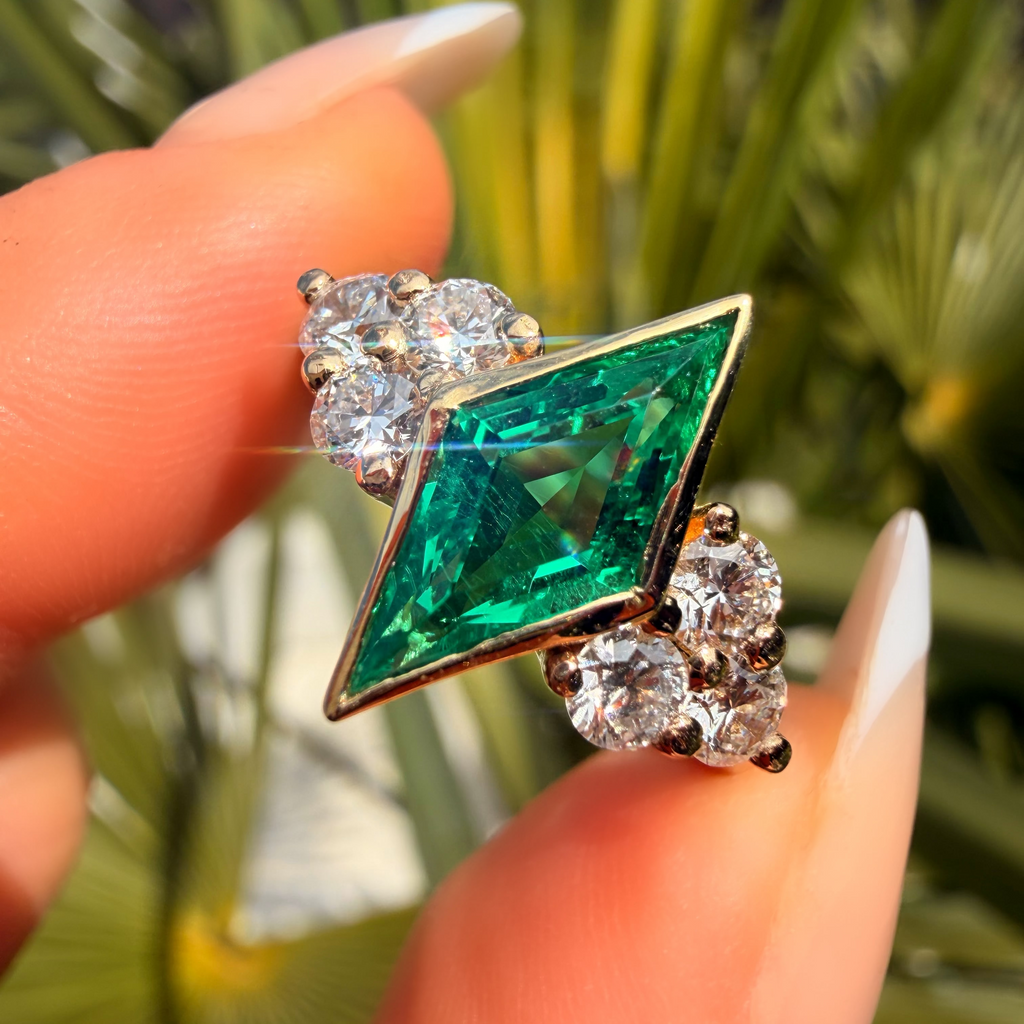 Emerald green gemstone ring with diamond accents held between fingers against a blurred natural background