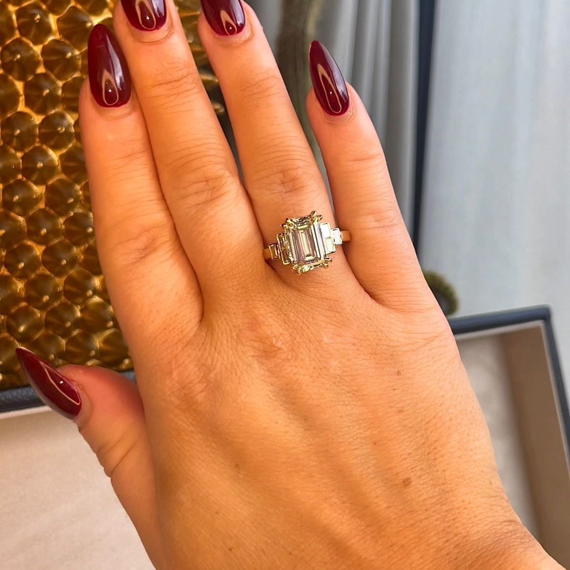 Hand with red nail polish wearing a large diamond ring against a textured wall.