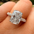 A close-up of a hand wearing a silver-toned ring with a large cushion-cut diamond and smaller diamonds along the band, set against a blurred outdoor background.