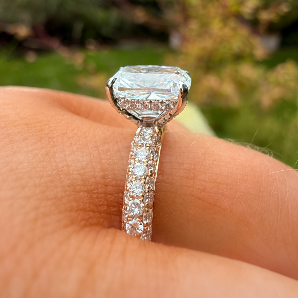 A close-up of a diamond ring with a large cushion-cut center stone and a pavé-set band, worn on a finger with a blurred outdoor garden background.