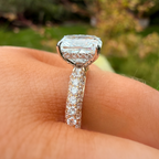 A close-up of a diamond ring with a large cushion-cut center stone and a pavé-set band, worn on a finger with a blurred outdoor garden background.