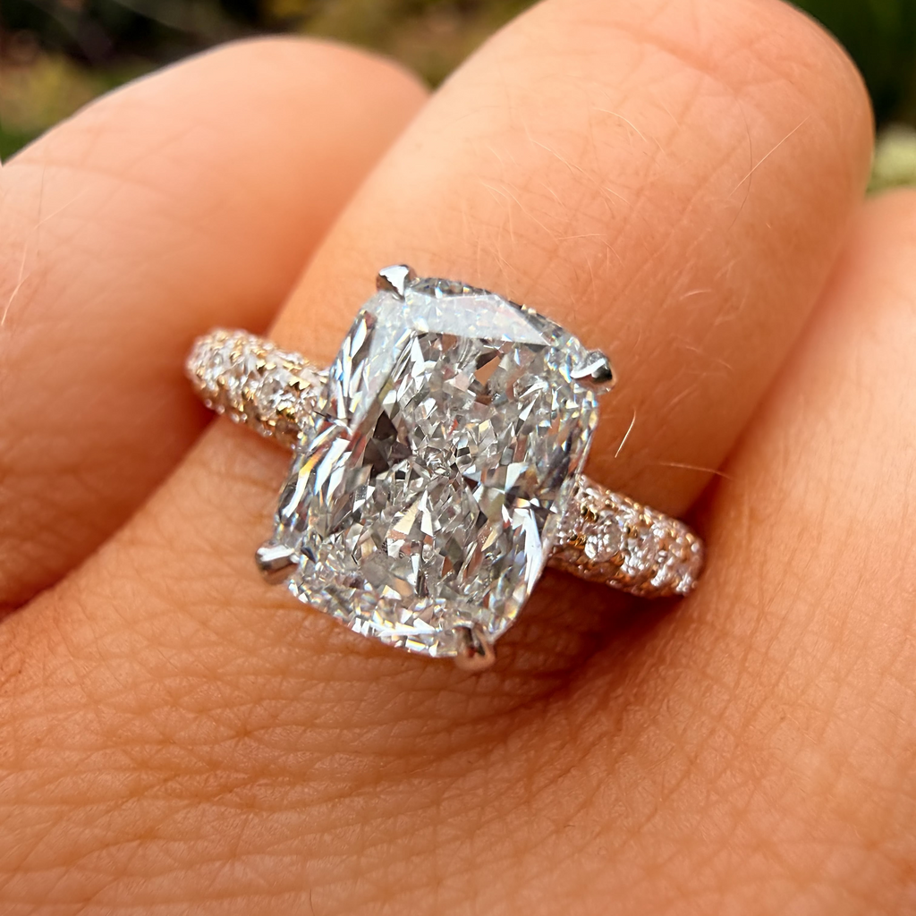 A close-up of a hand wearing a silver-toned ring with a large cushion-cut diamond and smaller diamonds along the band, set against a blurred outdoor background.
