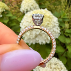 A close-up of a rose gold engagement ring with a large clear center stone and a band set with small clear stones, held by a hand with light pink nail polish against a blurred background of white hydrangea flowers and green foliage.