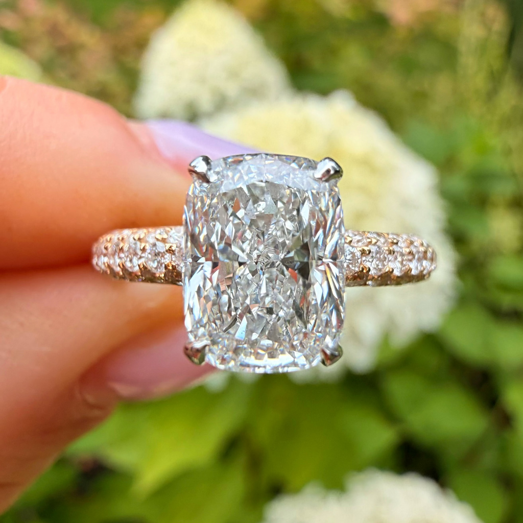 A close-up of a hand holding a large cushion-cut diamond engagement ring with a pavé band, set against a blurred background of green foliage and white flowers.