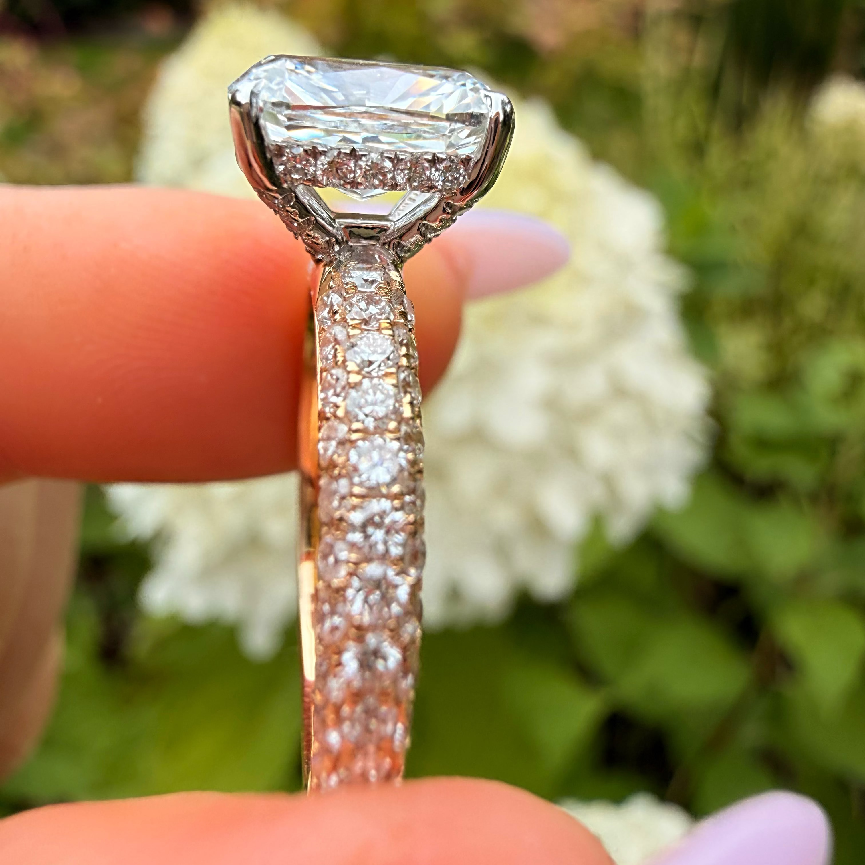 A close-up of a diamond ring held between fingers, featuring a large round-cut center stone and a pavé-set band, with a blurred garden background of green foliage and white flowers.