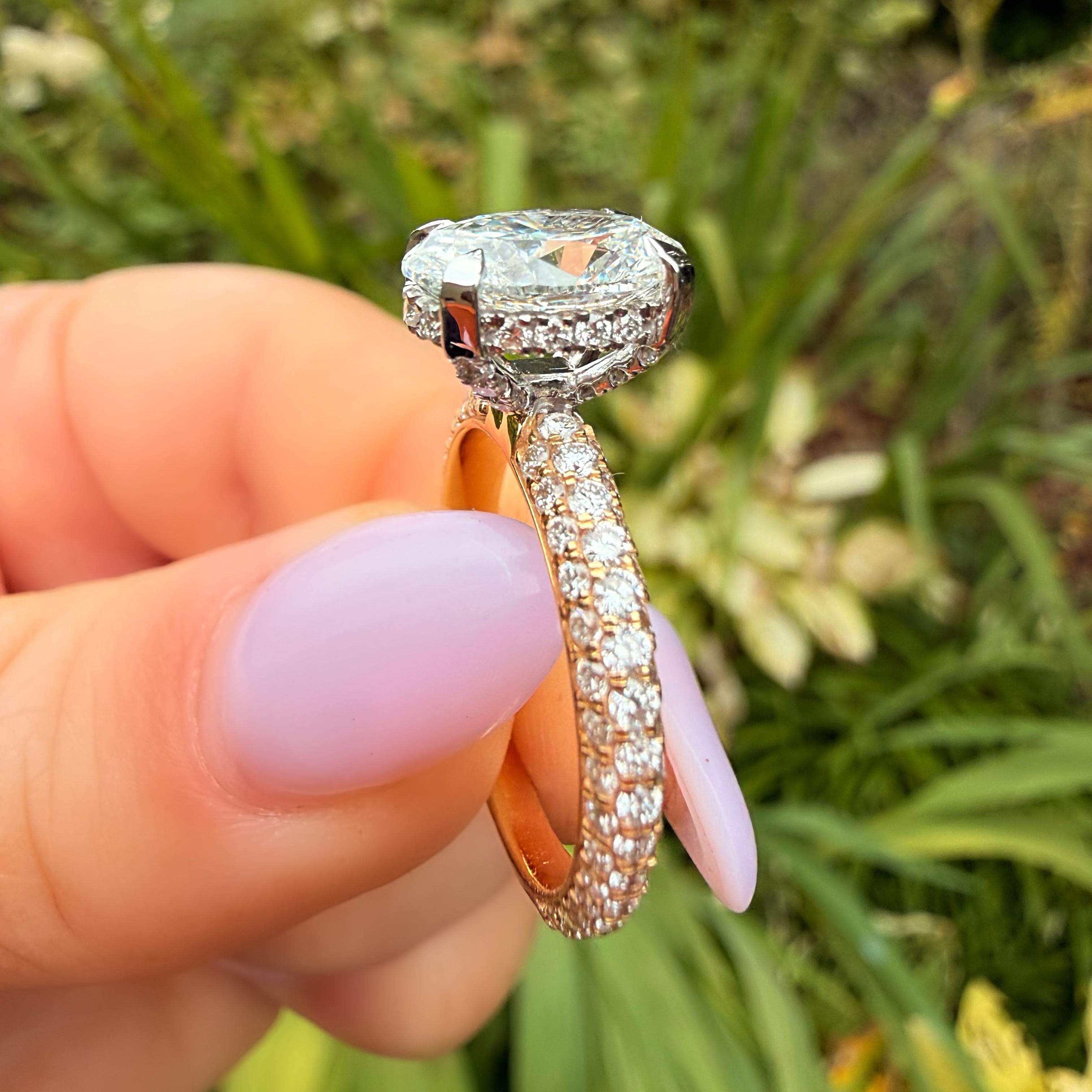 A close-up of a hand with light purple manicured nails holding a diamond ring outdoors with a blurred green garden background.